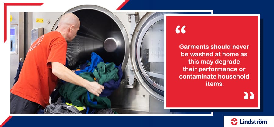 Worker loading PPE into industrial washing machine with garment care warning.