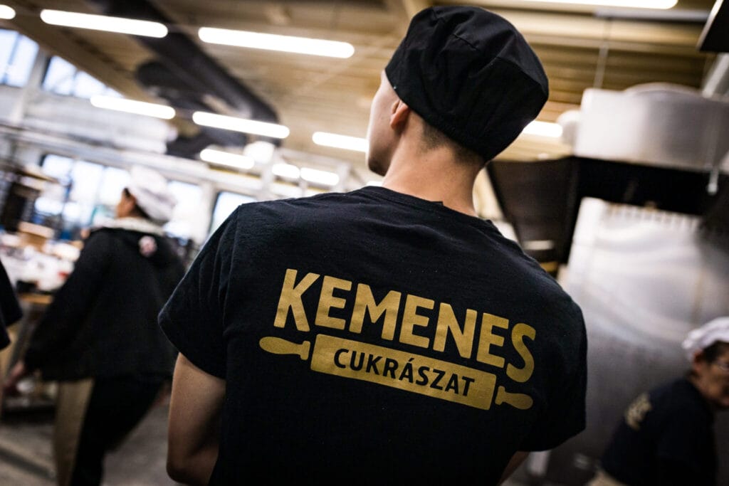 Kemenes konditerijos darbuotojas su juoda darbo apranga ir įmonės logotipu