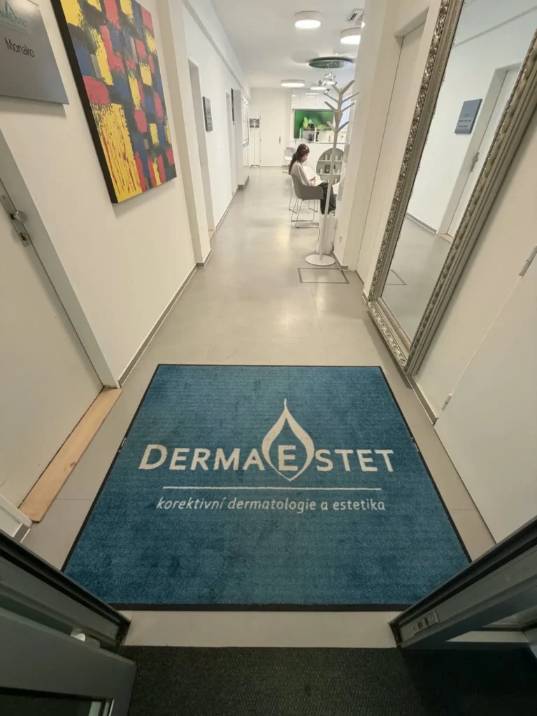 Modrá čisticí rohož s logem DERMAESTET u vstupu do dermatologické kliniky, která zachytává nečistoty a vlhkost z obuvi.