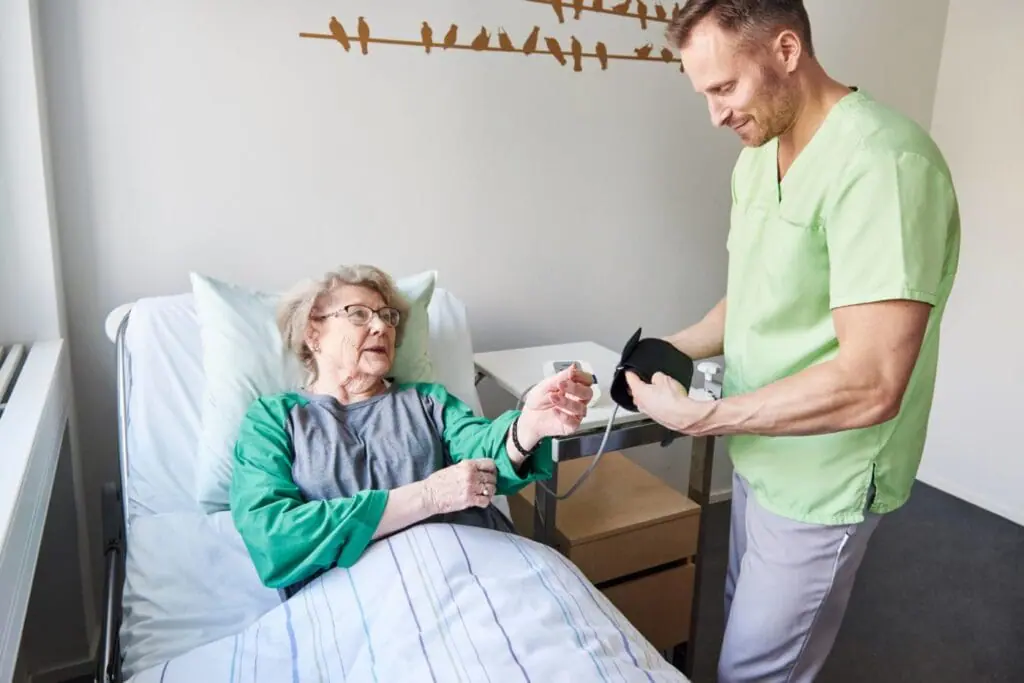 Zdravotník v zelené pracovní haleně měří krevní tlak seniorce na lůžku při poskytování zdravotní péče.