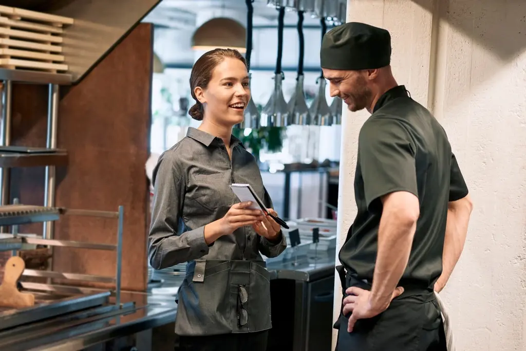 Restaurantleiterin spricht mit einem Koch in hygienischer Berufskleidung im Gastronomiebetrieb.
