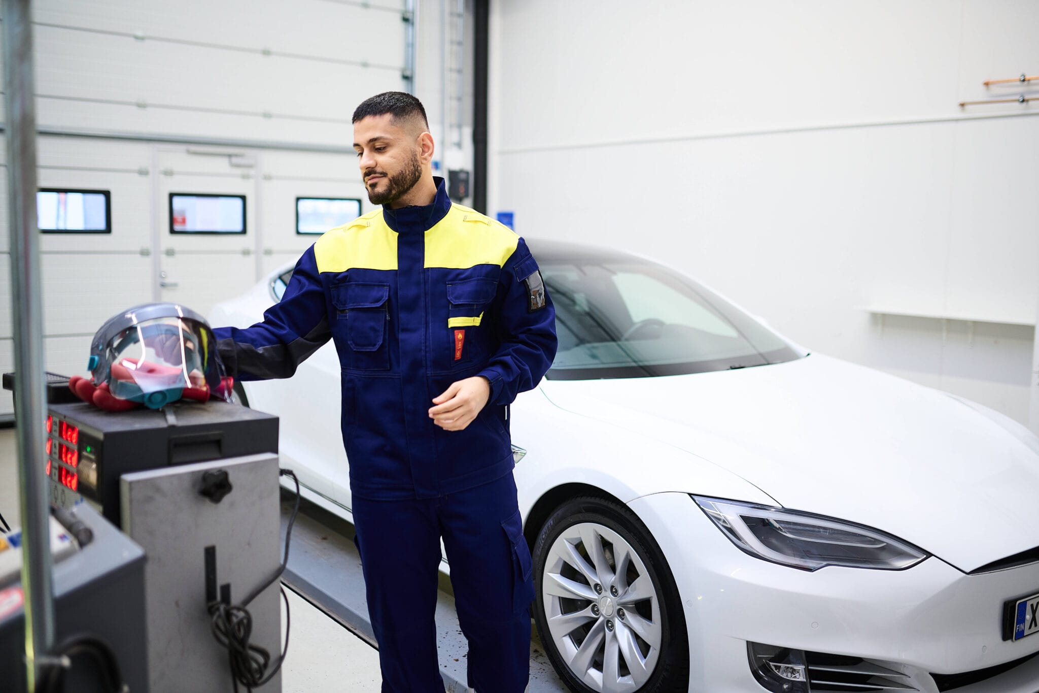 En servicetekniker i blå och gul arbetsuniform står i en ljus verkstad bredvid en vit elbil. Han håller i en skyddshjälm med visir som ligger på en testmaskin