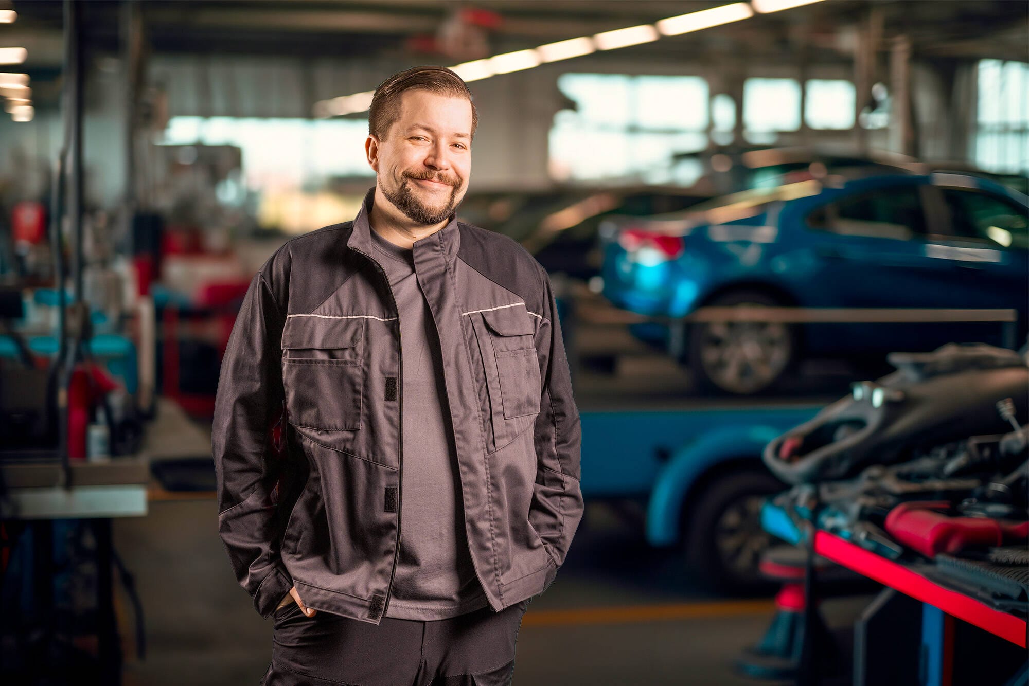 Porträtt av en leende servicetekniker i en grå arbetsjacka som står i en bilverkstad med blå bilar i bakgrunden
